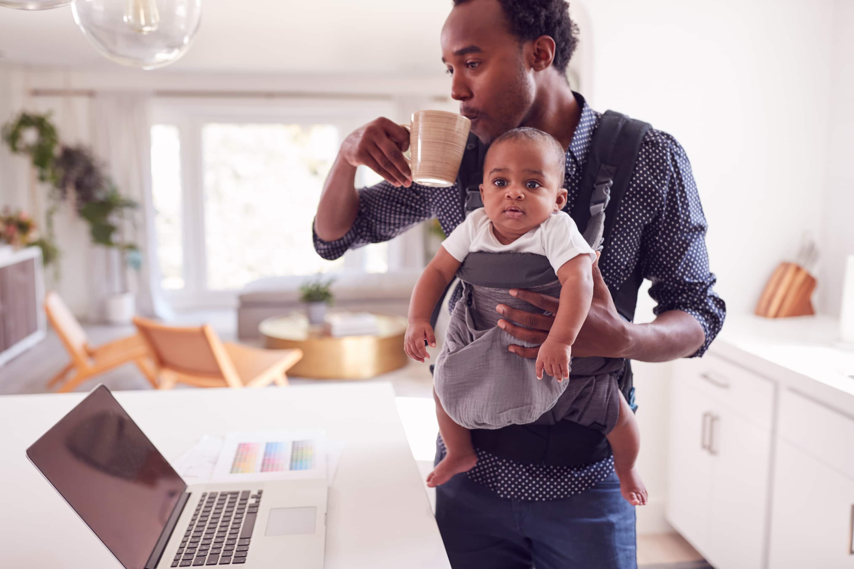 Father With Baby Daughter In Sling Multi Tasking Working From Home On Laptop